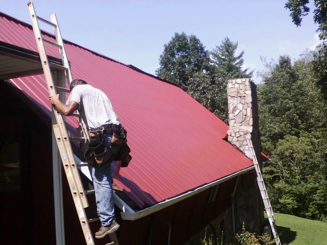 Roofer on a ladder beside a red metal roof and stone chimney — Soffit & Fascia Repair by Roofing Brothers in Marblehead